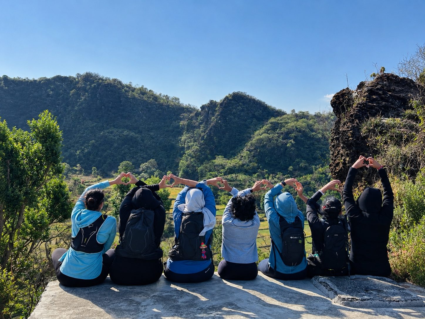 Seven women sitting on a ledge above green hills, their arms raised in heart shapes